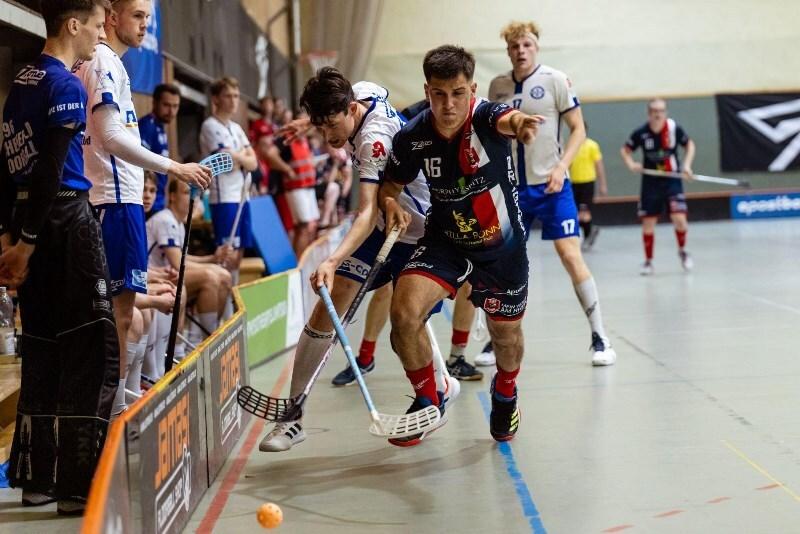 Zwei Spieler k&auml;mpfen um den Ball in einem Hallenboden-Hockeyspiel, w&auml;hrend Zuschauer auf der Bank zuschauen.