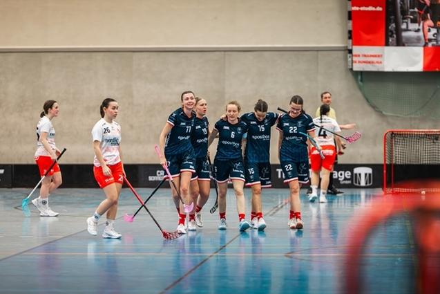 Sechs Frauen in Sportkleidung feiern auf dem Feld eines Unihockeyspiels in einer Halle.