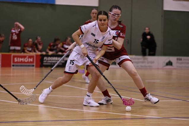Zwei Spielerinnen im Floorball k&auml;mpfen um den Ball, w&auml;hrend Zuschauer im Hintergrund zuschauen.