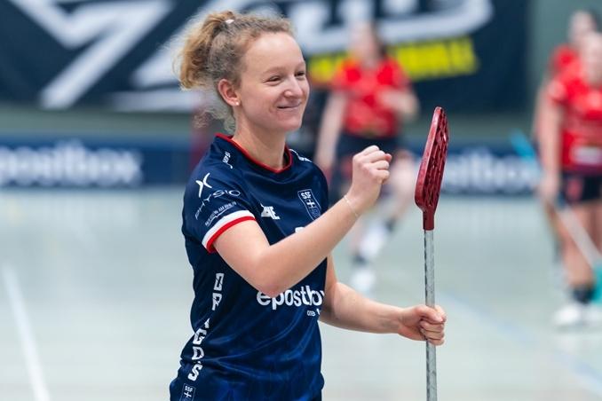 Freudige Spielerin mit Floorball-Schläger in sportlicher Kleidung, Hintergrund zeigt weitere Spielerinnen in Aktion.