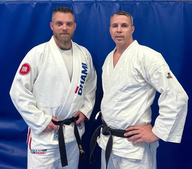 Zwei M&auml;nner in Judoanz&uuml;gen mit schwarzen G&uuml;rteln stehen vor einer blauen Wand.