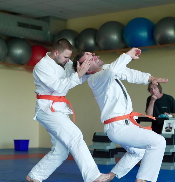 Zwei Judoka in der &Uuml;bung, einer h&auml;lt den anderen mit orangefarbenem G&uuml;rtel, im Hintergrund eine Trainingsumgebung.