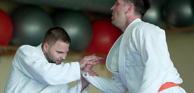 Zwei Judoka in wei&szlig;en Anz&uuml;gen &uuml;ben einen Griff in einem Trainingsraum mit Fitnessger&auml;ten im Hintergrund.