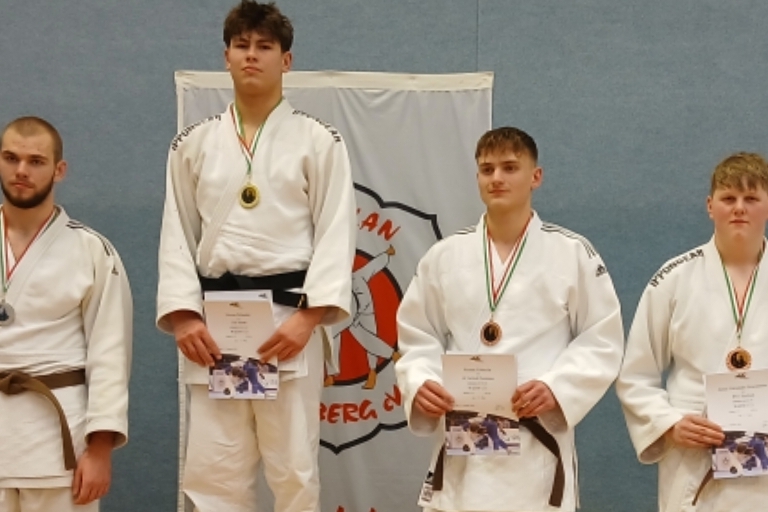 Gruppenfoto von Judokas in Mats, die Medaillen und Urkunden halten, vor einer Wand mit Judokas-Logo.
