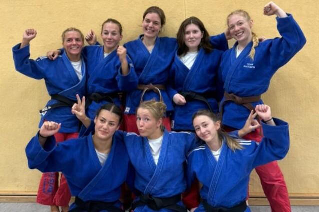 Gruppenbild von acht Judokas in blauen Anz&uuml;gen mit unterschiedlichen G&uuml;rtelfarben, alle l&auml;cheln und posieren.