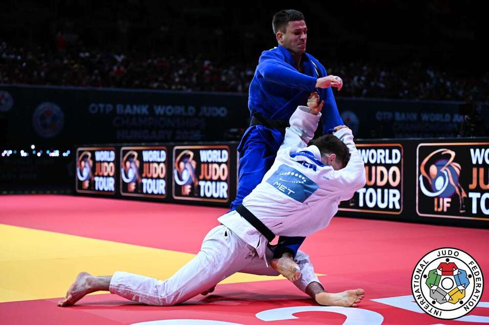 Zwei Judok&auml;mpfer in Aktion auf einer rot-gelben Matte w&auml;hrend eines Wettkampfs. Der K&auml;mpfer in Blau greift den Gegner in Wei&szlig; an.