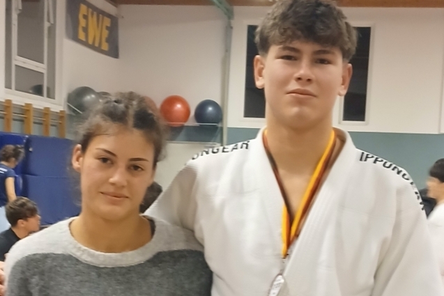 Zwei Jugendliche stehen in einem Trainingsraum, der Junge tr&auml;gt einen wei&szlig;en Judogi mit Medaille, die junge Frau in Sweatshirt.