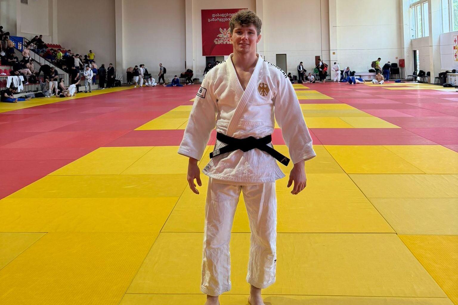 Junger Judoka in wei&szlig;em Gi mit schwarzem G&uuml;rtel auf einer bunten Judomatte, im Hintergrund sind andere K&auml;mpfer und Zuschauer.
