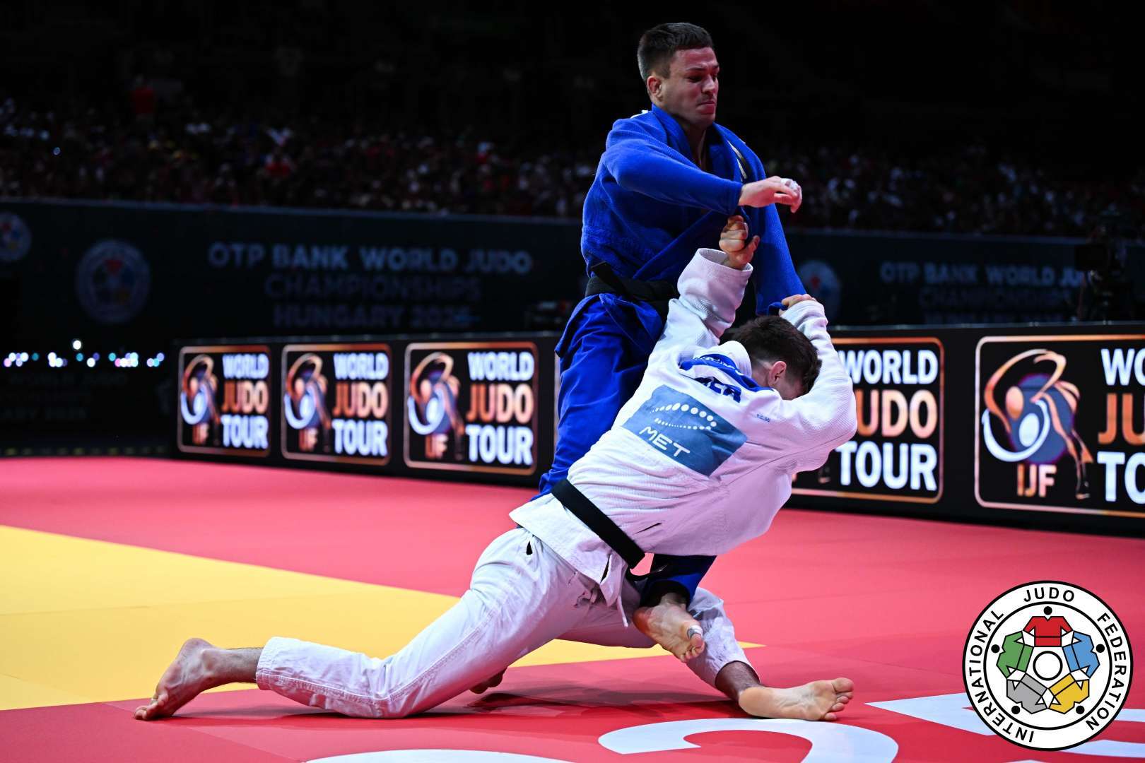 Zwei Judok&auml;mpfer in Aktion auf einer rot-gelben Matte w&auml;hrend eines Wettkampfs. Der K&auml;mpfer in Blau greift den Gegner in Wei&szlig; an.