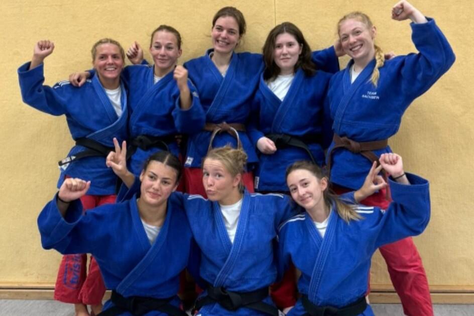 Gruppenbild von acht Judokas in blauen Anz&uuml;gen mit unterschiedlichen G&uuml;rtelfarben, alle l&auml;cheln und posieren.