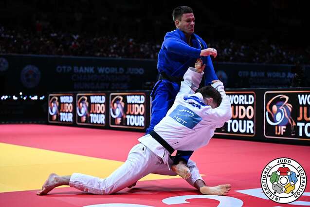 Zwei Judok&auml;mpfer in Aktion auf einer rot-gelben Matte w&auml;hrend eines Wettkampfs. Der K&auml;mpfer in Blau greift den Gegner in Wei&szlig; an.