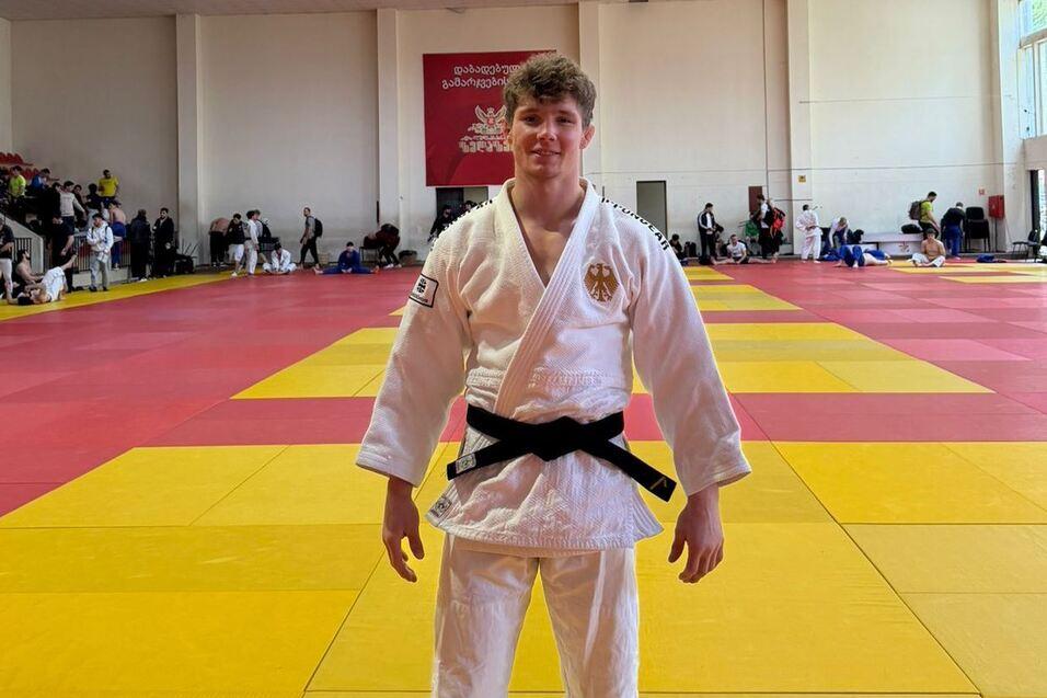 Junger Judoka in weißem Gi mit schwarzem Gürtel auf einer bunten Judomatte, im Hintergrund sind andere Kämpfer und Zuschauer.
