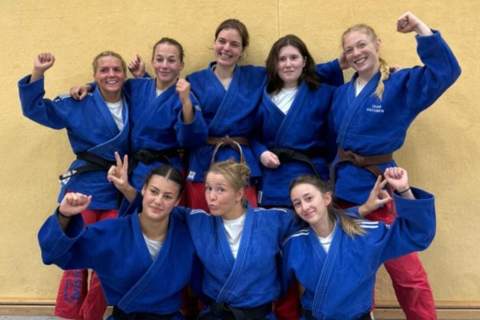 Gruppenbild von acht Judokas in blauen Anz&uuml;gen mit unterschiedlichen G&uuml;rtelfarben, alle l&auml;cheln und posieren.