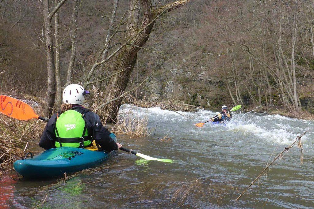Zwei Kajakfahrer auf einem rei&szlig;enden Fluss, einer paddelt in entgegenkommender Richtung. Ein Ufer mit B&auml;umen im Hintergrund.