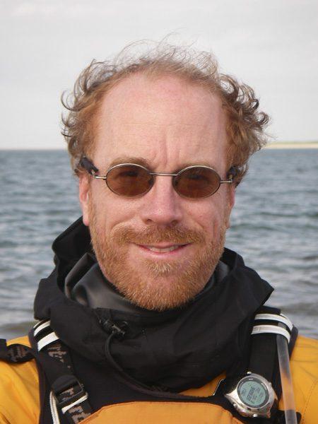 Portr&auml;t eines Mannes mit Brille und leichtem Bart, der in wetterfester Kleidung vor einer Wasseroberfl&auml;che steht.