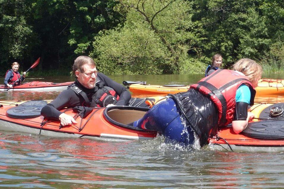 Zwei Personen in Kajaks auf einem Gew&auml;sser, eine Person klettert ins andere Kajak, umgeben von weiteren Paddlern.