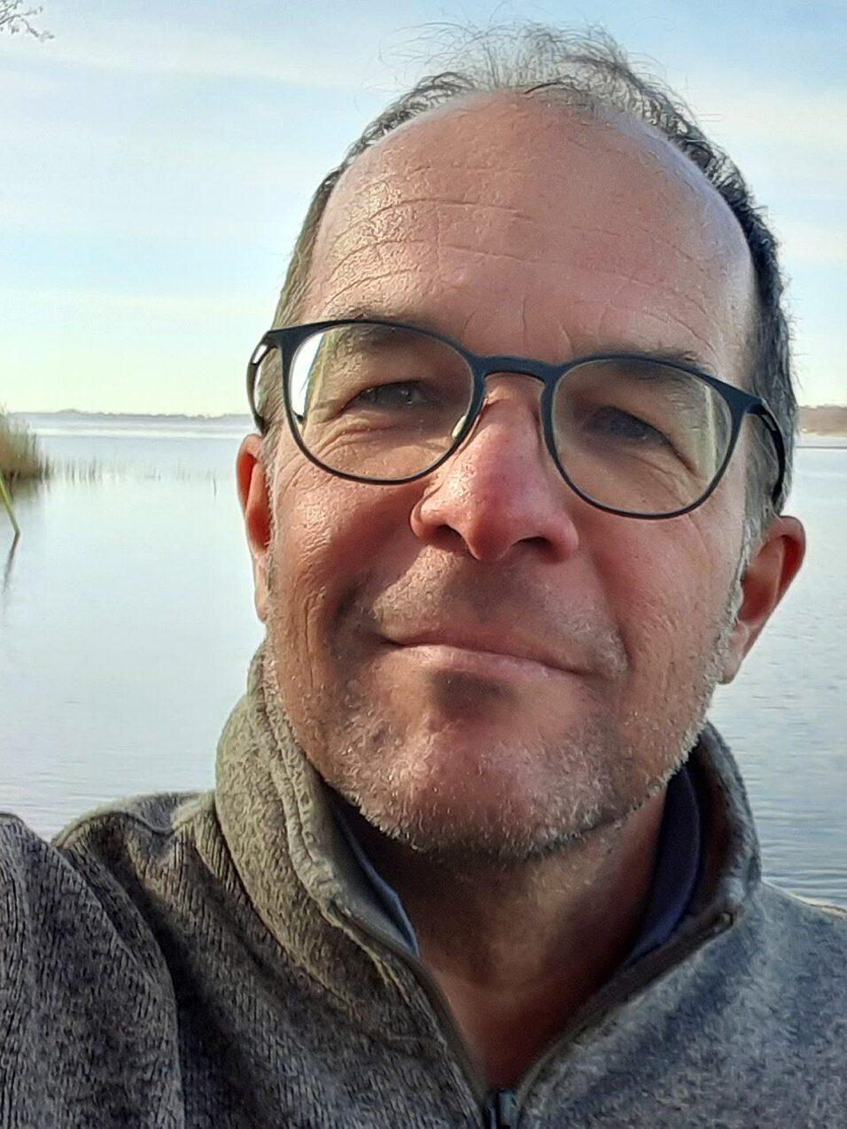 Selbstportr&auml;t eines Mannes mit Brille, l&auml;chelnd vor einem ruhigen See und leicht bew&ouml;lktem Himmel.