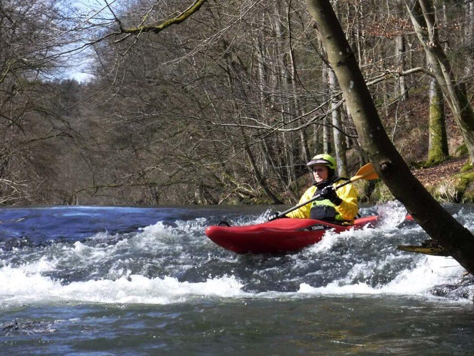 Kajakfahrer in gelber Kleidung paddelt durch einen reißenden Fluss, umgeben von Bäumen im Frühling.