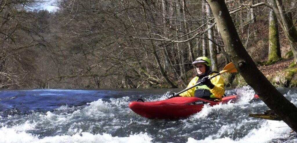 Kajakfahrer in gelber Kleidung paddelt durch einen rei&szlig;enden Fluss, umgeben von B&auml;umen im Fr&uuml;hling.