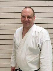 Mann in Karateanzug mit schwarzem G&uuml;rtel steht l&auml;chelnd vor einer Holzpaneelwand.