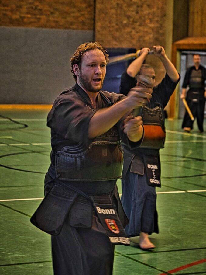 Zwei K&auml;mpfer in traditioneller Kendo-Ausr&uuml;stung &uuml;ben in einer Sporthalle miteinander.