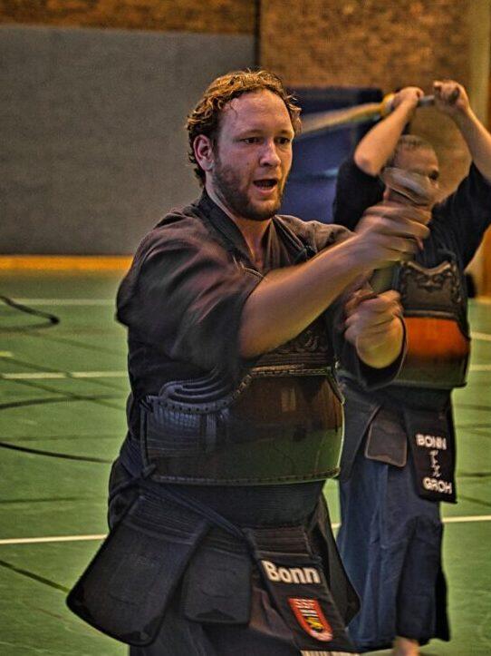 Zwei K&auml;mpfer in traditioneller Kendo-Ausr&uuml;stung &uuml;ben in einer Sporthalle miteinander.