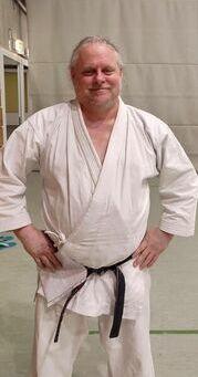Karateka in wei&szlig;em Gi und schwarzem G&uuml;rtel steht l&auml;chelnd in einer Sporthalle.