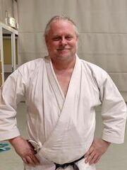 Karateka in wei&szlig;em Gi und schwarzem G&uuml;rtel steht l&auml;chelnd in einer Sporthalle.