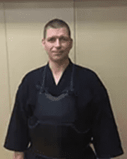 Mann in traditioneller japanischer Kampfsportkleidung mit Schutzanzug steht vor einer neutralen Wand.