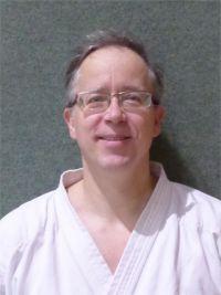 L&auml;chelnder Mann mit Brille, der einen wei&szlig;en Karateanzug tr&auml;gt, vor einer grau-gr&uuml;nen Wand.