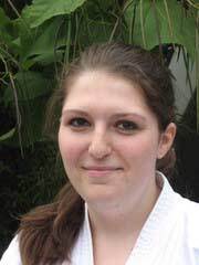 Portr&auml;t einer jungen Frau mit dunklen Haaren, die in einem wei&szlig;en Karateanzug vor einer gr&uuml;nen Pflanzenkulisse steht.