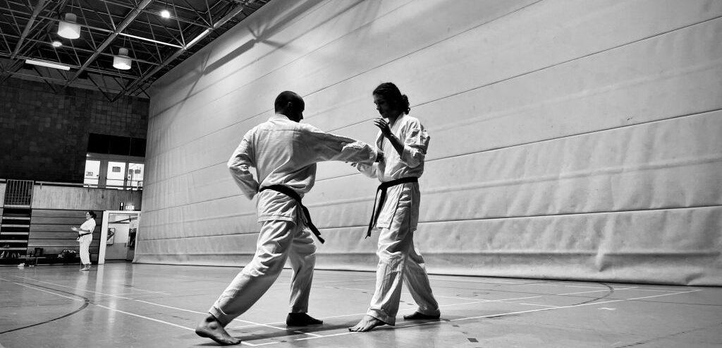 Zwei Kampfsportler in Karateanz&uuml;gen &uuml;ben Techniken in einer Sporthalle mit hellen W&auml;nden und Holzboden.