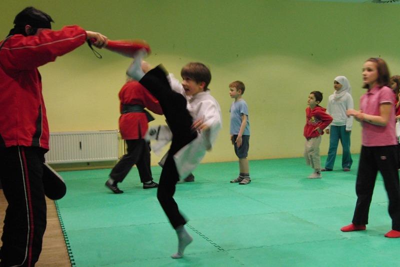 Kinder &uuml;ben Karate im Dojo; ein Junge f&uuml;hrt einen Kick gegen einen Trainer aus, w&auml;hrend andere zusehen.