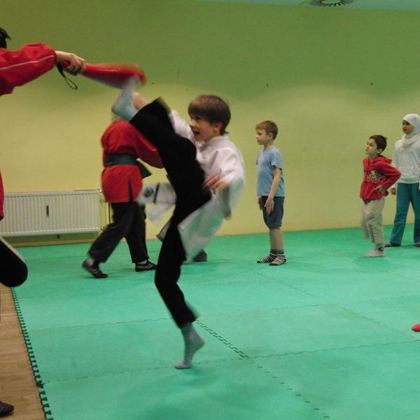 Kinder &uuml;ben Karate im Dojo; ein Junge f&uuml;hrt einen Kick gegen einen Trainer aus, w&auml;hrend andere zusehen.