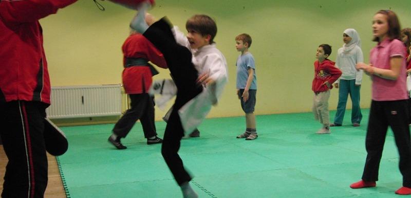 Kinder &uuml;ben Karate im Dojo; ein Junge f&uuml;hrt einen Kick gegen einen Trainer aus, w&auml;hrend andere zusehen.