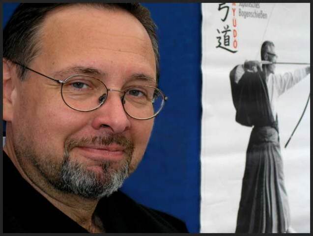 Mann mit Brille und Bart vor einem Poster mit einem Bogensch&uuml;tzen in traditioneller Kleidung.