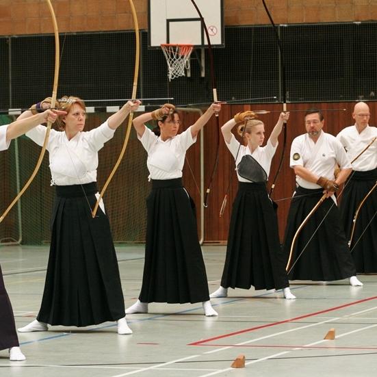 Sechs Personen in traditioneller Kleidung &uuml;ben Bogenschie&szlig;en in einer Sporthalle mit Basketballkorb.