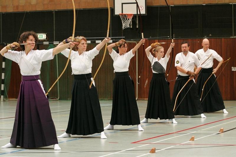 Sechs Personen in traditioneller Kleidung &uuml;ben Bogenschie&szlig;en in einer Sporthalle mit Basketballkorb.