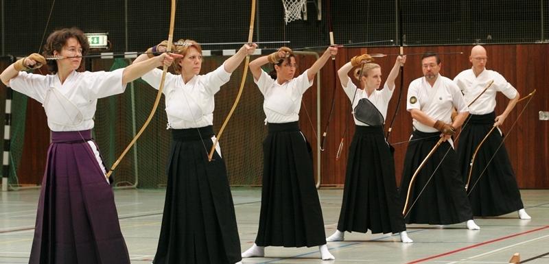 Sechs Personen in traditioneller Kleidung &uuml;ben Bogenschie&szlig;en in einer Sporthalle mit Basketballkorb.