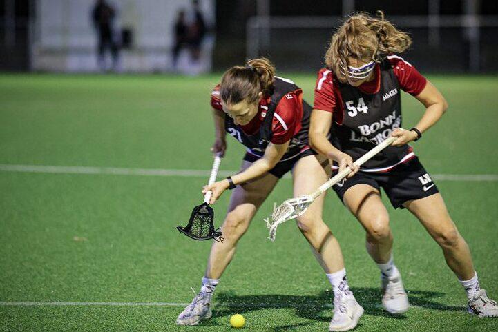 Zwei Lacrosse-Spielerinnen k&auml;mpfen um einen gelben Ball auf einem Kunstrasenfeld.