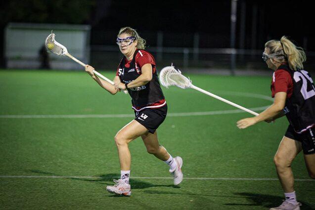 Zwei Lacrosse-Spielerinnen in sportlicher Kleidung trainieren auf einem Kunstrasenfeld unter Flutlicht.