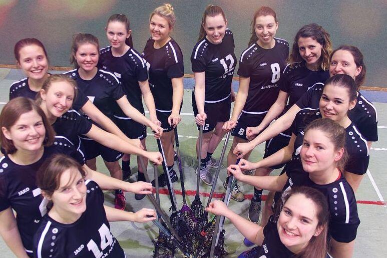 Gruppenaufnahme von 14 Frauen in schwarzen Trikots, die Lacrosse-Schl&auml;ger in der Mitte halten.