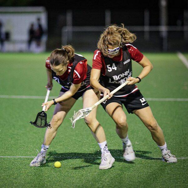 Zwei Lacrosse-Spielerinnen k&auml;mpfen um einen gelben Ball auf einem Kunstrasenfeld.