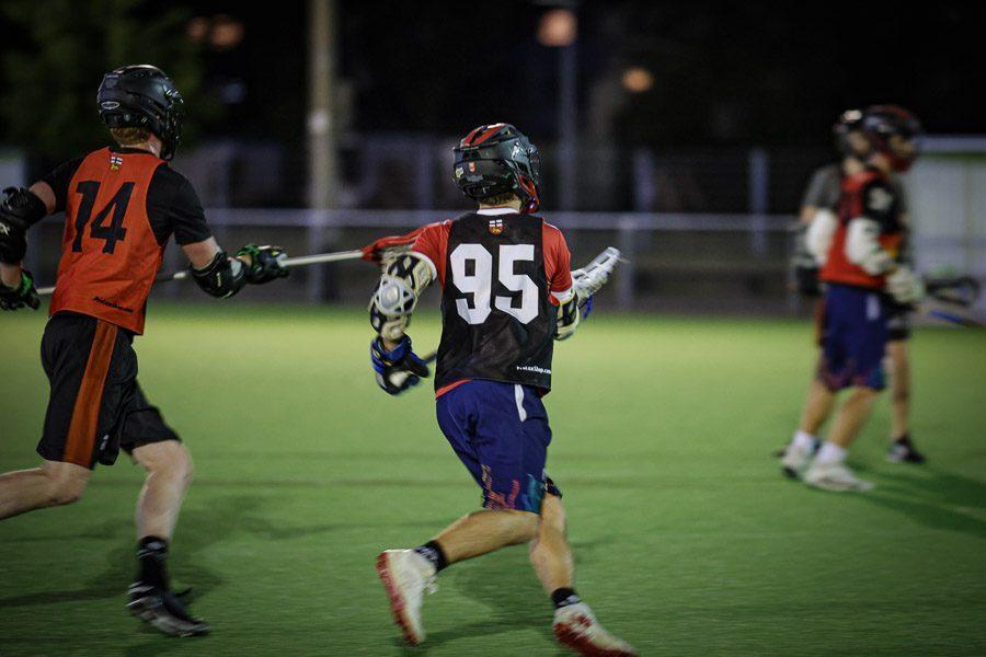 Zwei Lacrosse-Spieler in Sportkleidung bewegen sich auf einem beleuchteten Kunstrasenfeld bei Nacht.