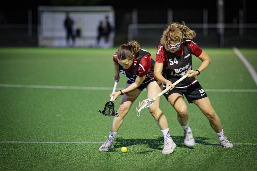 Zwei Lacrosse-Spielerinnen k&auml;mpfen um einen gelben Ball auf einem Kunstrasenfeld.