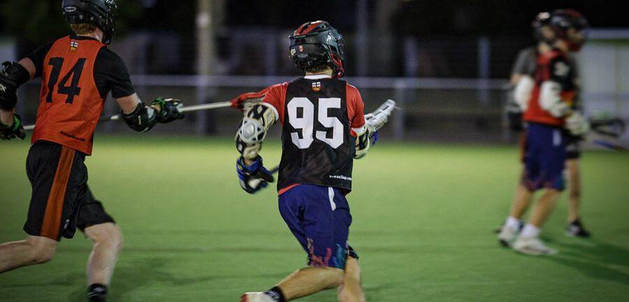 Zwei Lacrosse-Spieler in Sportkleidung bewegen sich auf einem beleuchteten Kunstrasenfeld bei Nacht.