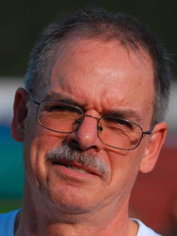 Nahaufnahme eines Mannes mit Brille und Schnurrbart, der direkt in die Kamera schaut. Hintergrund unscharf und farbig.