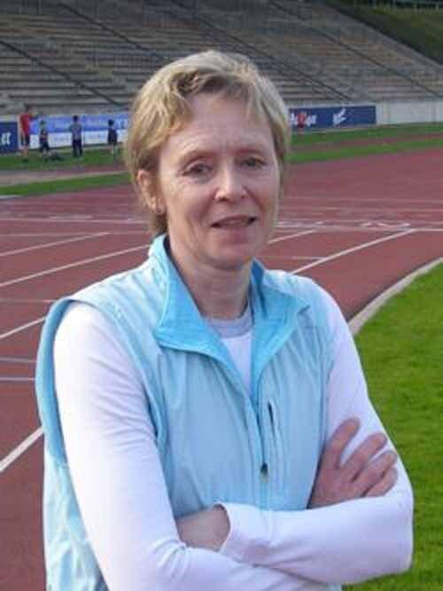 Frau mit kurzen blonden Haaren, in blauer Weste, steht l&auml;chelnd auf einer Laufbahn im Stadion.