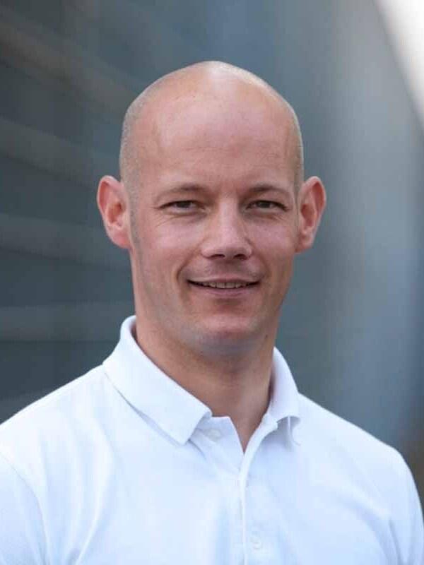 Portr&auml;t eines l&auml;chelnden Mannes mit kurzer, glatter Haarfrisur, in einem wei&szlig;en Polohemd vor einem unscharfen, grauen Hintergrund.