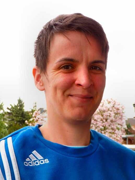 Person mit kurzem Haar in blauer Sportjacke steht vor bl&uuml;henden B&auml;umen und l&auml;chelt in die Kamera.